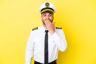 Arap uçak pilotu, sarı arka planda izole edilmiş mutlu ve gülümseyen ağızla elini kapatıyor.