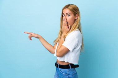 Genç Uruguaylı sarışın kadın mavi arka planda, sürpriz bir ifadeyle işaret ederken...