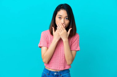 Mavi arka planda izole edilmiş genç Vietnamlı kadın elleriyle ağzını kapatıyor.