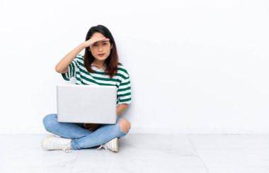 Diz üstü bilgisayarı olan genç Vietnamlı bir kadın beyaz duvarda izole edilmiş bir şekilde oturuyor. Bir şeye bakmak için çok uzaklara bakıyor.