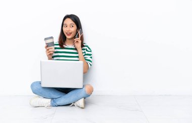 Yerde dizüstü bilgisayarı olan genç Vietnamlı bir kadın beyaz duvarda izole bir şekilde oturuyor. Elinde kahve ve cep telefonu var.