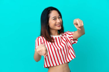 Genç Vietnamlı kadın mavi arka planda izole edilmiş, mutlu bir ifadeyle ön tarafı işaret ediyor.