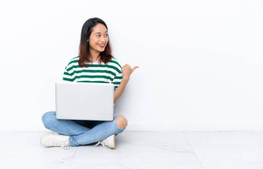 Yerde dizüstü bilgisayarı olan genç Vietnamlı bir kadın beyaz duvarda izole edilmiş bir şekilde oturuyor. Yan tarafı göstererek bir ürün sunuyor.