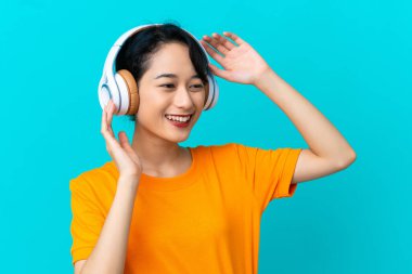 Genç Vietnamlı kadın mavi arka planda izole edilmiş müzik dinliyor ve şarkı söylüyor.