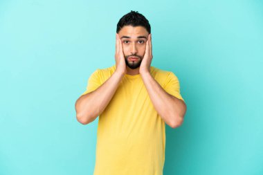 Genç Arap adam mavi arka planda izole edilmiş hayal kırıklığına uğramış ve kulaklarını kapatmış.