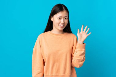 Genç Çinli kız mavi arka planda mutlu bir ifadeyle selamlıyor.