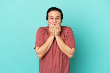 Kafkasyalı genç adam mavi arka planda izole edilmiş mutlu ve gülümseyen elleriyle ağzını kapatıyor.