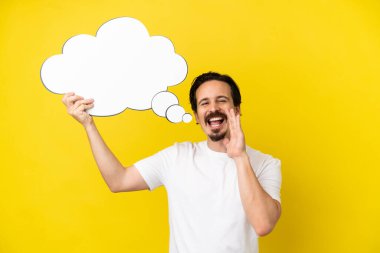 Young caucasian man isolated on yellow background holding a thinking speech bubble and shouting