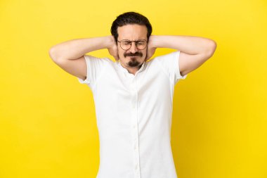 Sarı arka planda izole edilmiş genç beyaz adam hayal kırıklığına uğramış ve kulaklarını kapatmış.