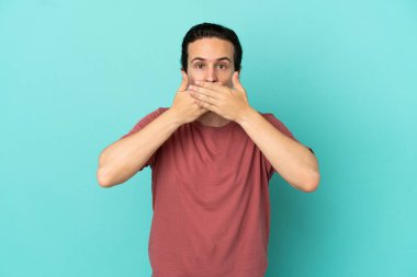 Mavi arka planda izole edilmiş genç beyaz adam elleriyle ağzını kapatıyor.