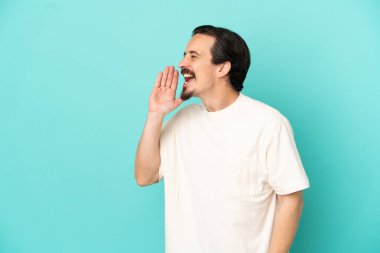 Mavi arka planda izole edilmiş genç beyaz adam ağzı sonuna kadar açık bağırıyor.