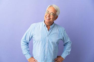 Middle age Brazilian man isolated on purple background posing with arms at hip and smiling