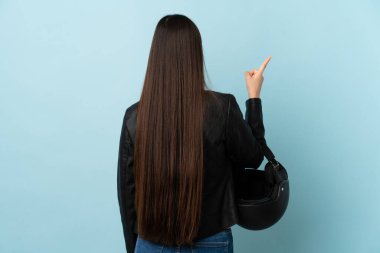 Chinese woman holding a motorcycle helmet over isolated blue background pointing back with the index finger