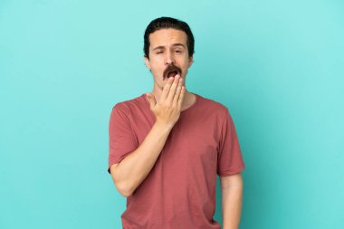 Kafkasyalı genç adam mavi arka planda izole edilmiş esniyor ve geniş açık ağzını eliyle kapatıyor.