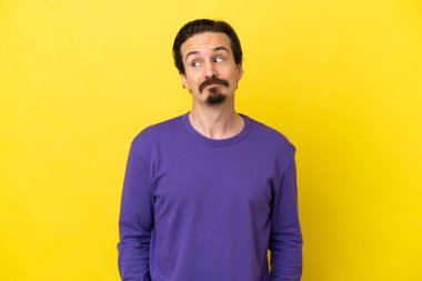 Young caucasian man isolated on yellow background making doubts gesture looking side