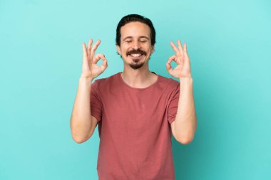 Zen pozisyonunda mavi arka planda izole edilmiş genç beyaz bir adam.
