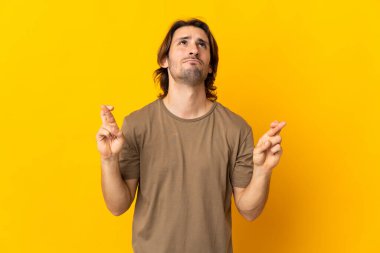 Sarı arka planda izole edilmiş genç ve yakışıklı bir adam parmaklarını çapraz yapıp en iyisini diliyor.