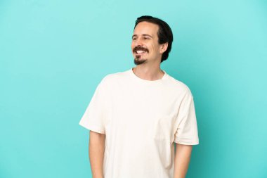 Kafkasyalı genç adam mavi arka planda izole edilmiş, yana bakıyor ve gülümsüyor.