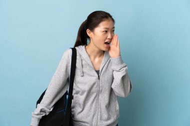 İzole edilmiş mavi arka planda spor çantalı genç Çinli kadın ağzı sonuna kadar açık bağırıyor.