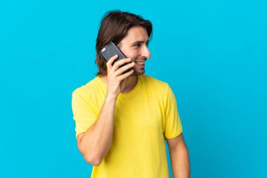 Genç ve yakışıklı bir adam mavi arka planda izole edilmiş, cep telefonuyla sohbet ediyor.