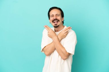 Mavi arka planda izole edilmiş genç beyaz adam gülümsüyor ve zafer işareti gösteriyor.