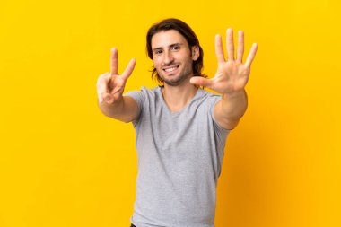 Sarı arka planda izole edilmiş genç yakışıklı yediyi parmaklarıyla sayıyor