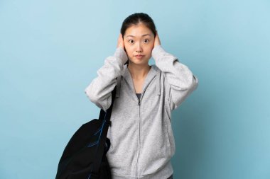 İzole edilmiş mavi arka planda spor çantalı genç Çinli kadın hayal kırıklığına uğramış ve kulaklarını kapatmış.
