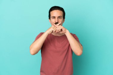Mavi arka planda izole edilmiş genç beyaz bir adam sessizlik işareti gösteriyor.