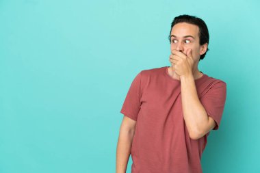 Mavi arka planda izole edilmiş genç beyaz adam yan tarafa bakarken sürpriz jest yapıyor.