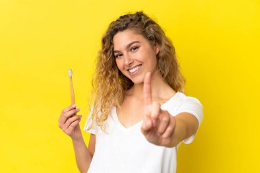 Sarı arka planda dişlerini fırçalayan genç beyaz kadın parmağını kaldırıyor ve gösteriyor.