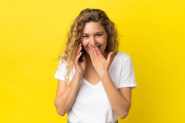 Sarı arka planda izole edilmiş cep telefonu kullanan genç sarışın kadın mutlu ve ağzı kapalı gülümsüyor.