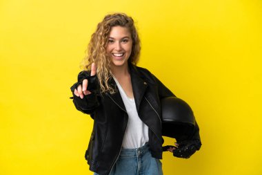 Sarı arka planda izole edilmiş motosiklet kasklı genç sarışın kadın parmağını kaldırıyor ve gösteriyor.