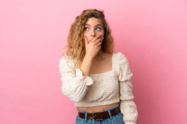 Pembe arka planda izole edilmiş genç sarışın kadın yan tarafa bakarken sürpriz jest yapıyor.