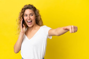 Sarı arka planda izole edilmiş cep telefonu kullanan genç sarışın kadın işaret veriyor.