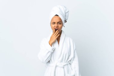 Bornozlu genç bir kadın izole edilmiş beyaz arka planda esniyor ve geniş bir ağzı eliyle kapatıyor.