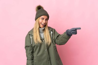 Kış şapkalı genç bir kadın pembe arka planda izole edilmiş bir şekilde parmak ucunda bir ürün sunuyor.