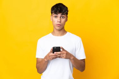 Sarı arka plan izole edilmiş genç adam kameraya bakarken şaşırmış bir ifadeyle cep telefonunu kullanıyor.