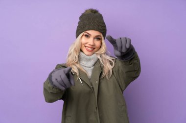 Kışlık şapkalı, mor arka plandaki genç sarışın kız gülümserken seni işaret ediyor.