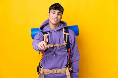 Sarı arka plan üzerinde büyük bir sırt çantası olan genç dağcı parmağını kaldırıyor ve gösteriyor.