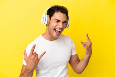 Kafkasyalı yakışıklı adam izole edilmiş sarı arka planda müzik dinliyor ve rock hareketleri yapıyor.