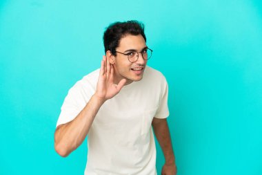 Kafkasyalı yakışıklı adam mavi arka planda elini kulağına koyarak bir şeyler dinliyor.