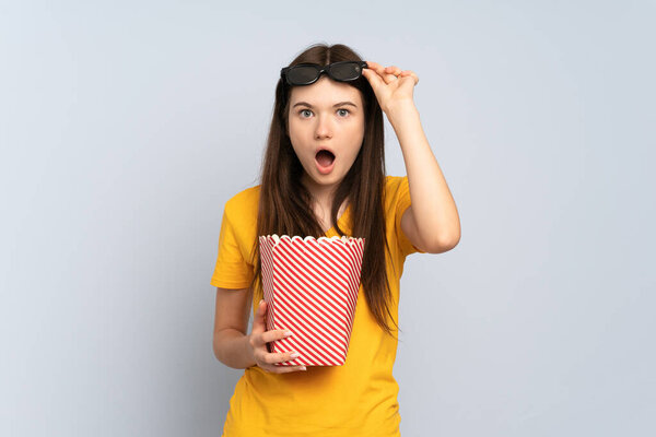 Young Ukrainian girl isolated on white background surprised with 3d glasses and holding a big bucket of popcorns