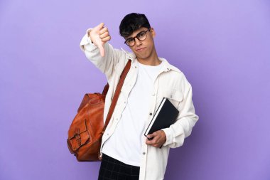 İzole edilmiş mor arka planda genç bir öğrenci, olumsuz ifadeyle baş parmağını aşağı gösteriyor.
