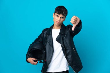 İzole edilmiş mavi arka plan üzerinde motosiklet kaskı olan genç bir adam negatif ifadeyle baş parmağını aşağı gösteriyor.