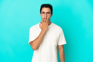 İzole edilmiş mavi arka planda, ağzı elle kaplayan beyaz, yakışıklı bir adam.
