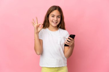 Pembe arka plan üzerinde izole edilmiş bir çocuk cep telefonu kullanıyor ve tamam işareti yapıyor