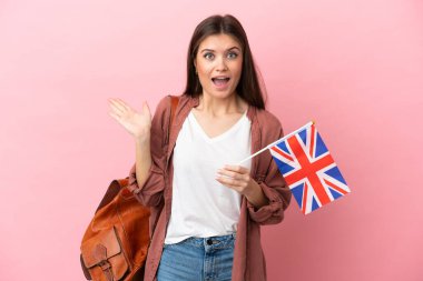 Elinde Birleşik Krallık bayrağı olan beyaz bir kadın pembe arka planda şok olmuş bir yüz ifadesiyle izole edilmiş.