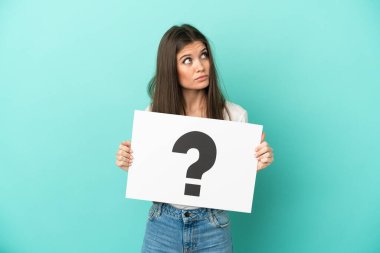 Kafkasyalı genç bir kadın mavi arka planda izole edilmiş. Elinde soru işareti olan bir levha var ve düşünüyor.