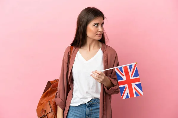 Young caucasian woman holding an United Kingdom flag isolated on pink background looking to the side