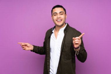 Asyalı yakışıklı adam mavi arka planda izole edilmiş, parmak uçlarını işaret ediyor ve mutlu.